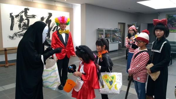 様々な仮装に扮した子供たちがカオナシからお菓子をもらっている様子を頭に紙で作られたカラフルな色の帽子、赤いタキシードに金色の大きな蝶ネクタイを付けて見守っている市長の写真