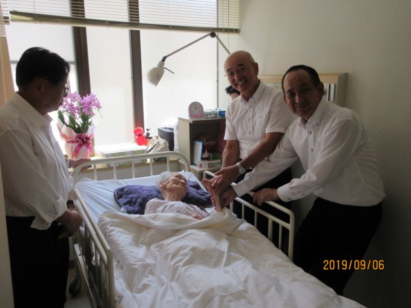 ベッドで横たわるご高齢の女性の周りを数名が心配そうにお祝いしている写真