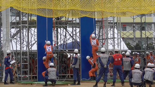 上から吊るされたロープを2名の隊員が登ろうとしていて、周りの人たちがその様子を見ている写真