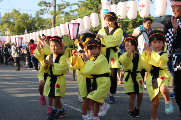 黄色い法被を羽織り頭にねじりハチマキをつけた園児たちが道路を歩きながら踊っている写真