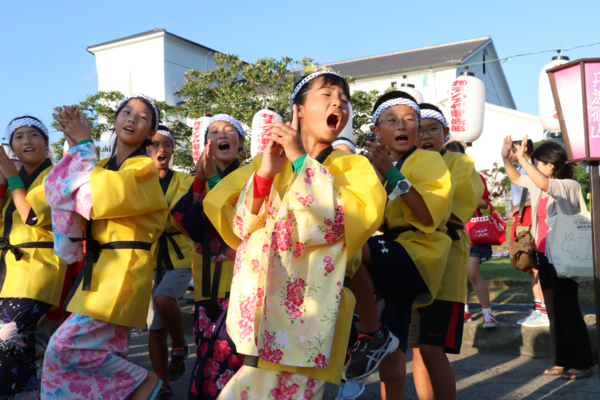 浴衣を着た女の子が法被を羽織り歌いながら踊って、その後ろで男の子たちも一緒に踊っている写真