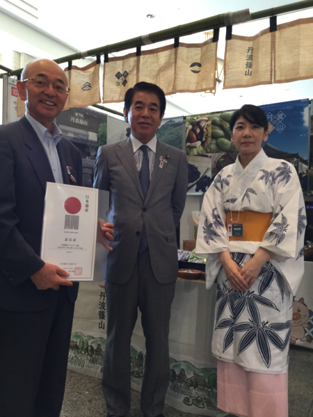 市長が認定証を手に持っていて、その横にスーツ姿の男性と着物姿の女性が並んでいる写真