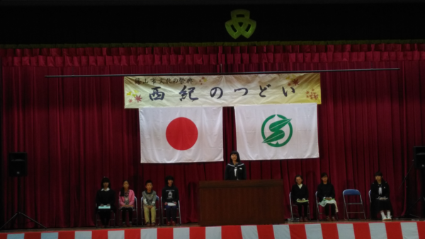 壇上で女の子が話をして、その後ろで椅子に座り子供達が話を聞いている写真