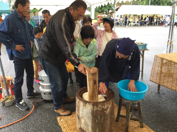 女性の方が水が入っている桶に手を入れて餅に水を付け、女の子と黒い服を着た男性が一緒に杵をついている。その後ろに順番待ちで並んでいる人々が写っている写真