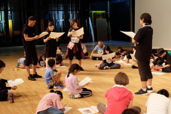 座って皆さん台本を持ち、指導者と3名が立って台本をみている写真