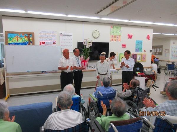 大勢のご高齢の方がひとりの男性に向かって拍手をしている写真