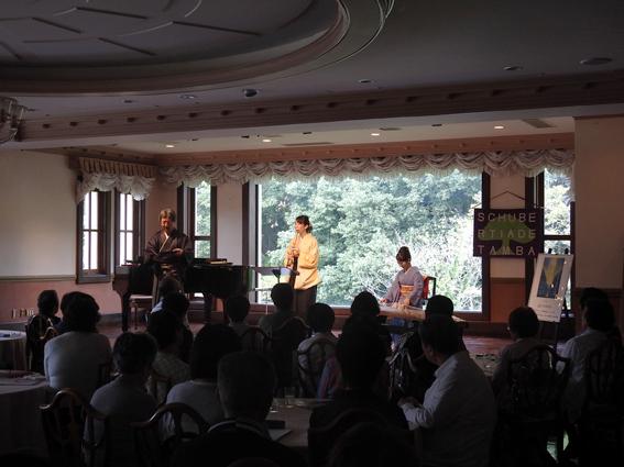 照明が落ちた室内にて、袴を着た井本 蝶山さん、畑 儀文さん、着物を着た女性が楽器を演奏している様子を観客が聞いている写真