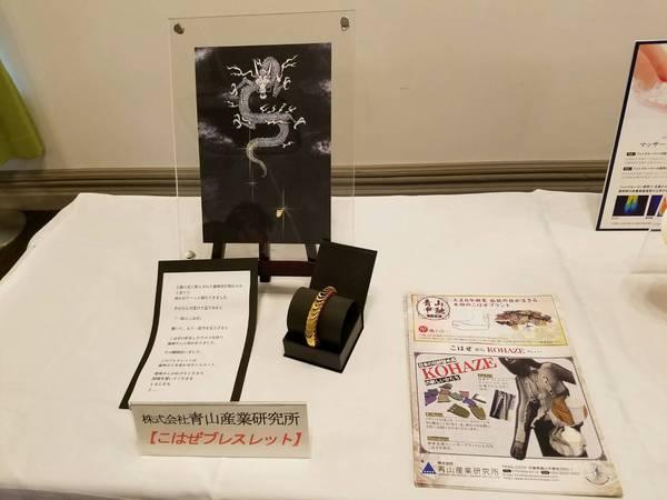 黒い箱の中の筒に巻かれているこはぜをつかった金色のブレスレットがパンフレットやパネルと一緒にテーブルに展示されている写真