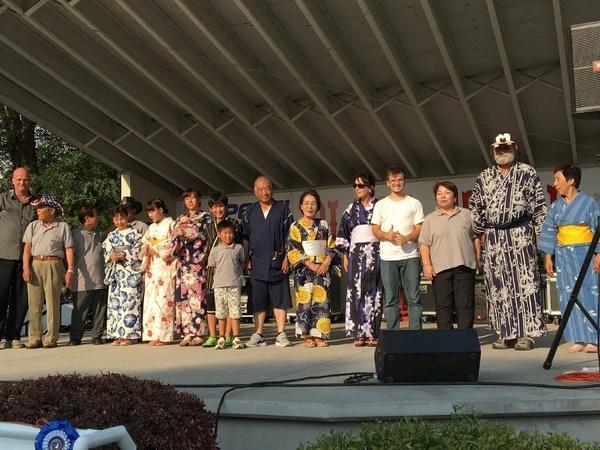 屋根のついた舞台の上で浴衣をきた訪問団の人がいたり浴衣姿の市民がいたりしてみんなで並んで写した集合写真