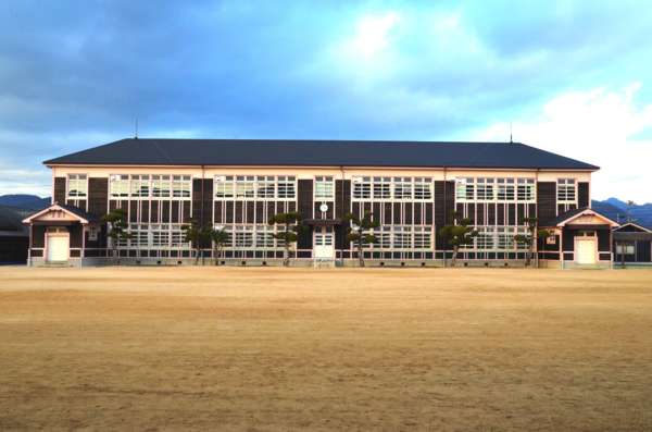 芝生の奥に木造の茶色い横長の建物が建っている写真