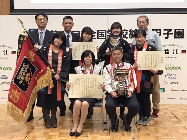 舞台上でトロフィーや賞状、優勝旗をもちメダルをかけ笑顔で整列している男性女性九名