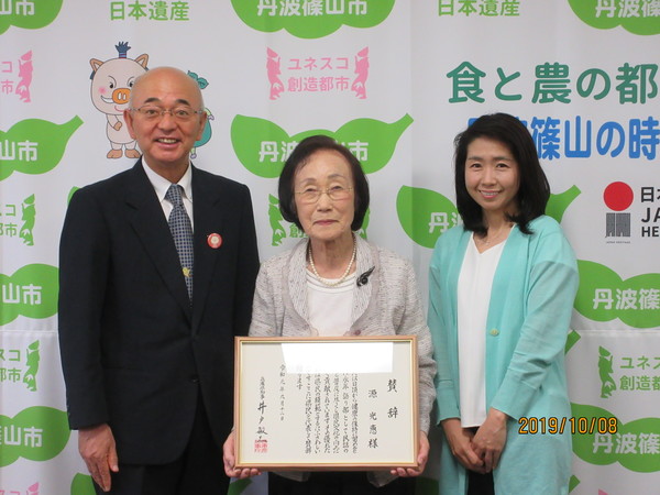 丹波篠山市と書かれた壁の前で市長と賞状をもった源さんと源家お嫁の貴美さんが笑顔で立っている写真