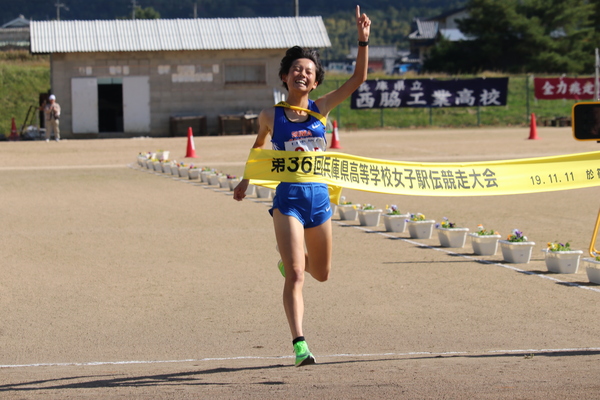 青いユニフォームを着た女子高校生が一番と指さしながら笑顔でゴールテープをきっている瞬間の写真