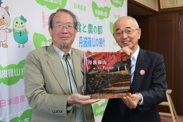 丹波篠山市と書かれた壁の前で写真家三木さんと市長が丹波篠山の写真集をもって笑顔で立っている写真