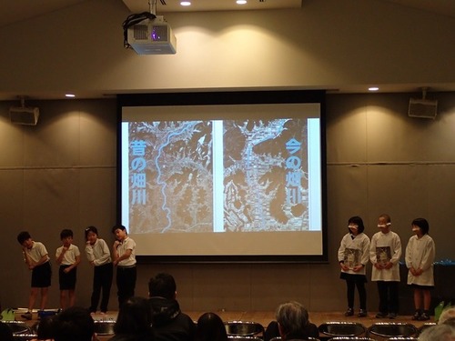 丹波篠山いきもの48フォーラムでの児童たちの発表の様子を写した写真。舞台には児童が上がっており、スクリーンには上から見た今と昔の畑川の写真が写されている。