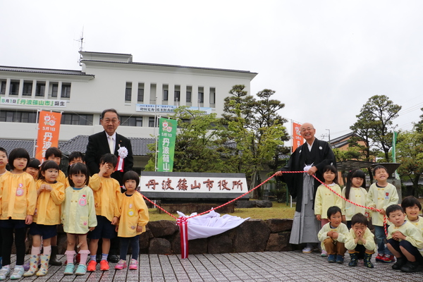丹波篠山市役所と書かれた銘板の除幕を行う市長と市議会議長。一緒に紅白のひもを引っ張る黄色い制服を着た子どもたち