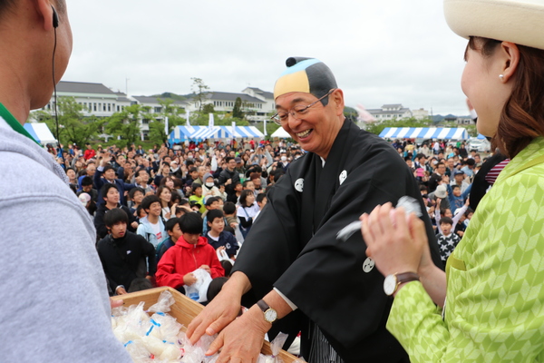 広場に集まる大勢の方々へお配りするお餅を持って、渡そうとしている市長と観光大使の女性