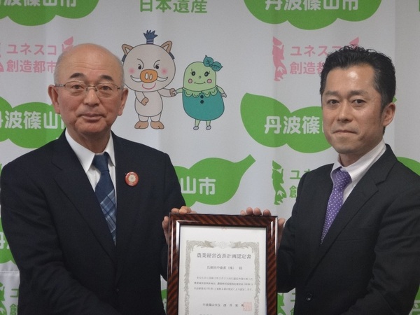 丹波篠山市と書かれた壁の前で、市長と田中さんが額に入った認定証を一緒に持っている写真