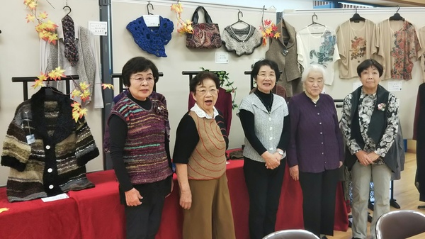 壁に飾られた衣服や作品の前で女性五名が笑って並んで立っている写真