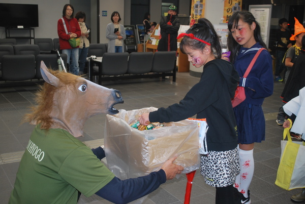 顔にペイントした女の子が列を作って馬のお面をかぶった大人からお菓子をもらっている様子
