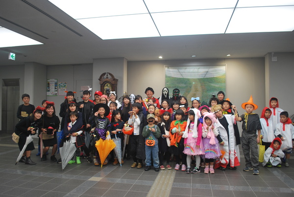 市民ホールでハロウィーン衣装に着飾った子どもたちや大人が笑って並んで立っている写真
