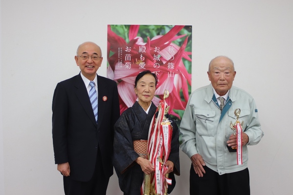 菊花展のポスター前に市長、青山賞受賞の西田さん、稲山さんがトロフィーをそれぞれ持って立っておられる写真
