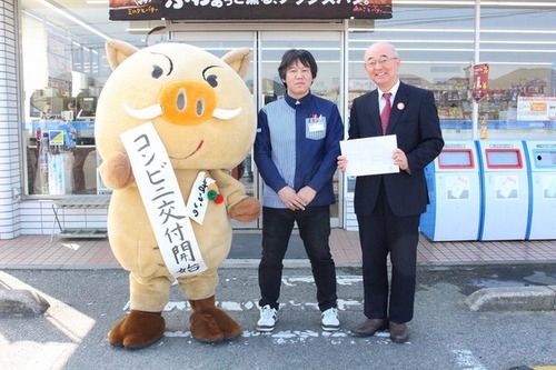 コンビニ交付開始と書かれた紙を持ったまるいのと市長が一緒に映っている写真。コンビニの前で撮られている。