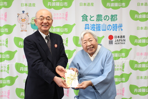 丹波篠山市と書かれた壁の前で、藤木さんから寄付金を両手で差しだして、両手で受け取る市長の写真