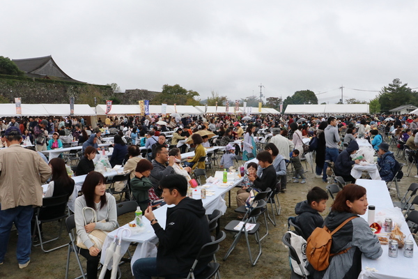 三の丸広場に広げられた椅子とテーブルで購入した特産物をおいしそうに食べる観光客のにぎわった様子