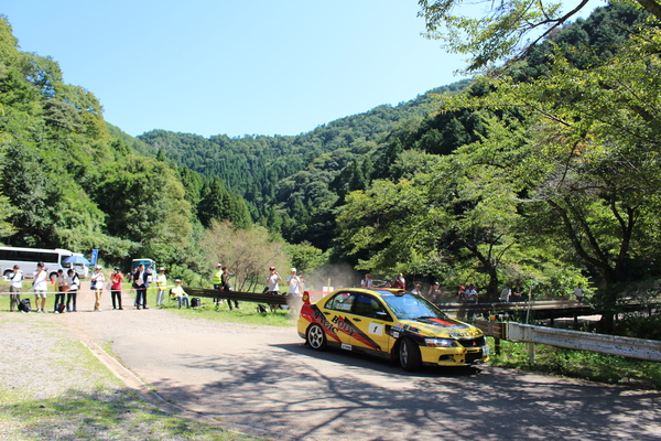 自然の中を数人のギャラリーが見守る中、黄色い車が走っている様子の写真