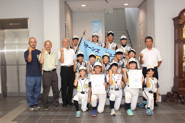 市民ホールのエレベーター横でユニフォームを着た子どもたちと市長、男性の方々がガッツポーズをしながら笑顔で立っている写真