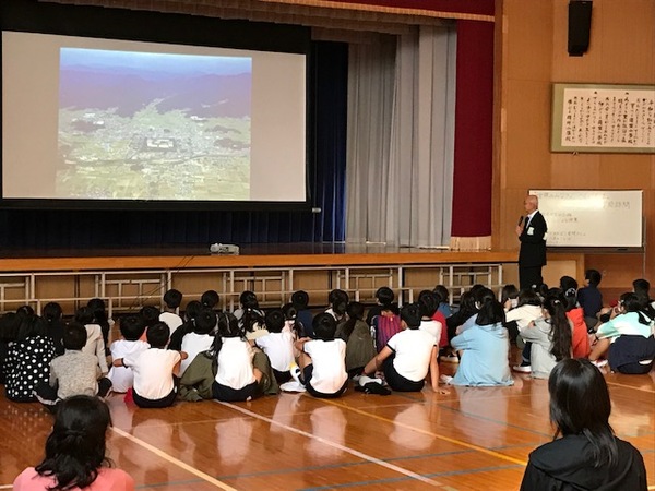 大きなスクリーンで投影しながらマイクをもって説明する市長とそれを床に座って聞く小学生たちの写真