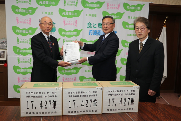 丹波篠山市と書かれた壁の前で、市長が西潟さん、小山さんより署名を受け取っている写真