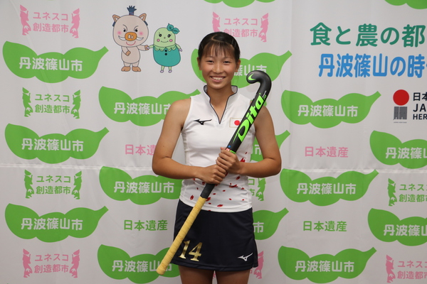 丹波篠山市と書かれた壁の前で、中井さんがホッケースティックをもって笑顔で立っている写真