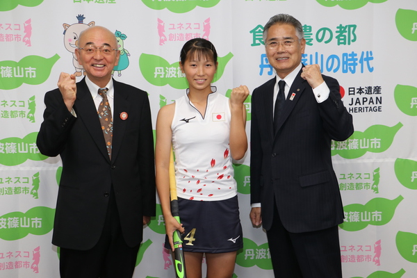 丹波篠山市と書かれた壁の前で市長、ホッケースティックを持った中井さん、前川教育長がガッツポーズをして笑顔で立っている写真