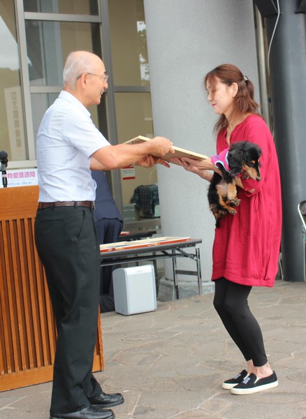 洋服を着た長寿犬が飼い主に抱かれて市長から表彰状を受け取っている写真