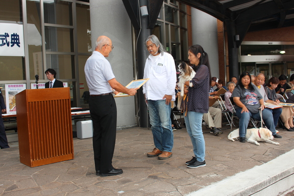 黄色いリードをつけた長寿犬と飼い主夫妻が市長から表彰状を受け取っている写真