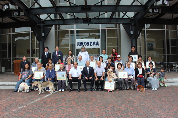 長寿犬表彰式典の幕を背景に表彰された犬と飼い主の方々が表彰状と一緒にうつる写真