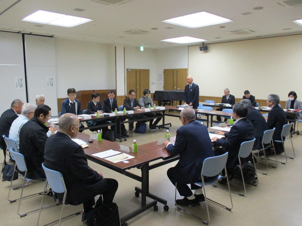 部屋の中央に机を円にして複数の人が椅子にかけて、一名立って話す男性の話を聞いている様子