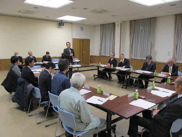 部屋の中央に机を円にして複数の人が椅子にかけて、一名立って話す男性の話を聞いている様子