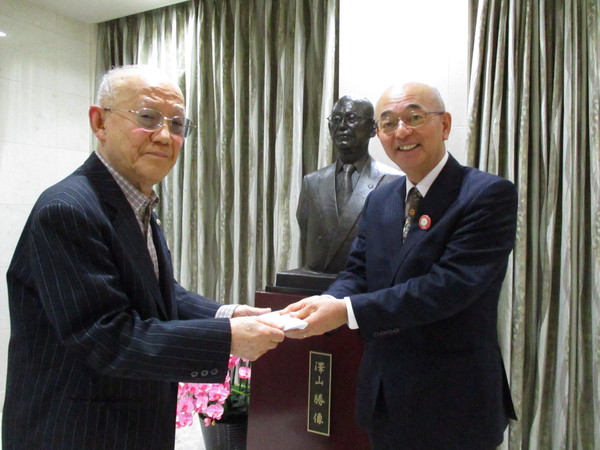 大きなカーテンと銅像の前で寄附金を手渡すスーツ姿の澤山氏と受け取る市長