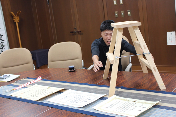 園田さんが四方転び踏み台と賞状、メダルを前に説明している様子の写真