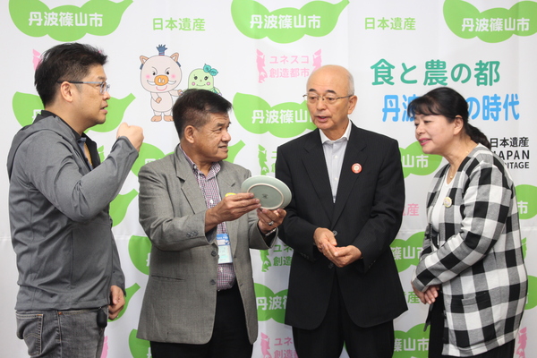 丹波篠山市と書かれた壁の前で市長とチェンマイの方々がお皿をもって談笑している写真