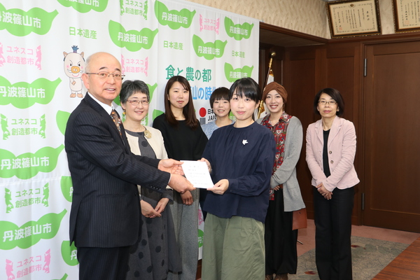丹波篠山市と書かれた壁の前で、女性から提案書をさせしだされ受け取る市長、それを見守る五名の女性の写真