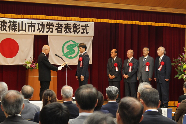 功労者表彰式典舞台上で市長から表彰を受けている五名の男性が並んでいる様子の写真