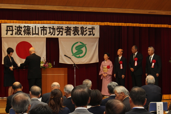 功労者表彰式典舞台上で市長から表彰を受けている四名の男性女性が並んでいる写真
