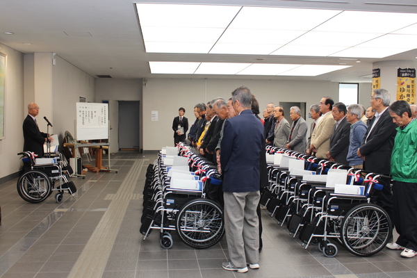 市民ホールにて車いす二十六台が並べられその間に男性が並んでマイクの前で話す市長の言葉を聞いている様子