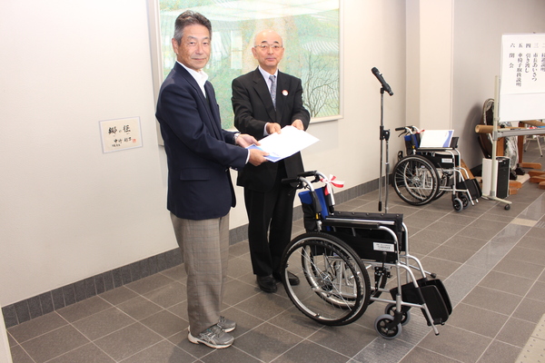 市民ホールにて寄贈された車いすを前にスーツ姿の男性から市長が書類を受け取っている写真