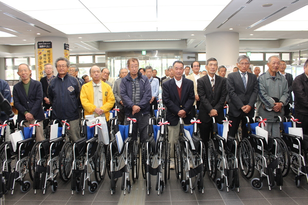 市民ホールにて贈られた車いすを前に男性が整列して並んで立っている様子