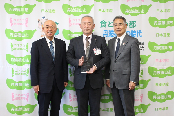 丹波篠山市と書かれた壁の前で市長、ガラスのトロフィーを持つ小林課長、前川教育長が笑顔で立っている写真